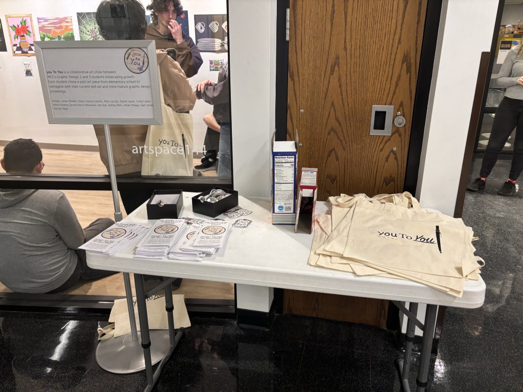 photo of a merchandise table for an art exhibition with paper brochures, buttons, fruit snacks, and tote bags