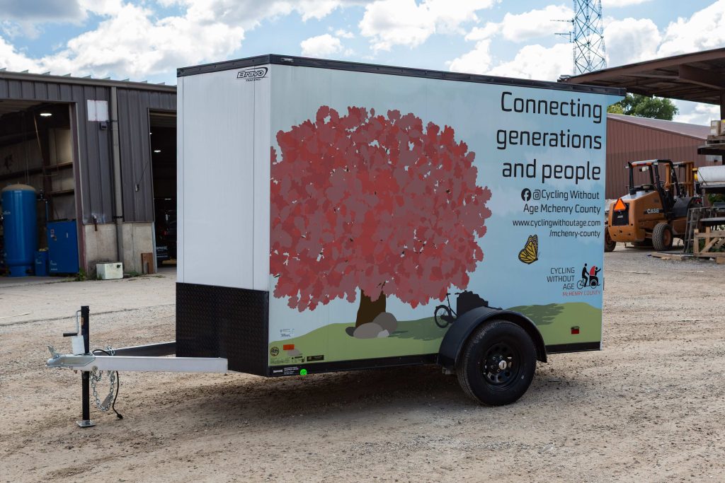 photo of a trailer with a wrap illustration depicting a tree with red leaves and a butterfly advertising cycling without age that reads "connecting generations and people"