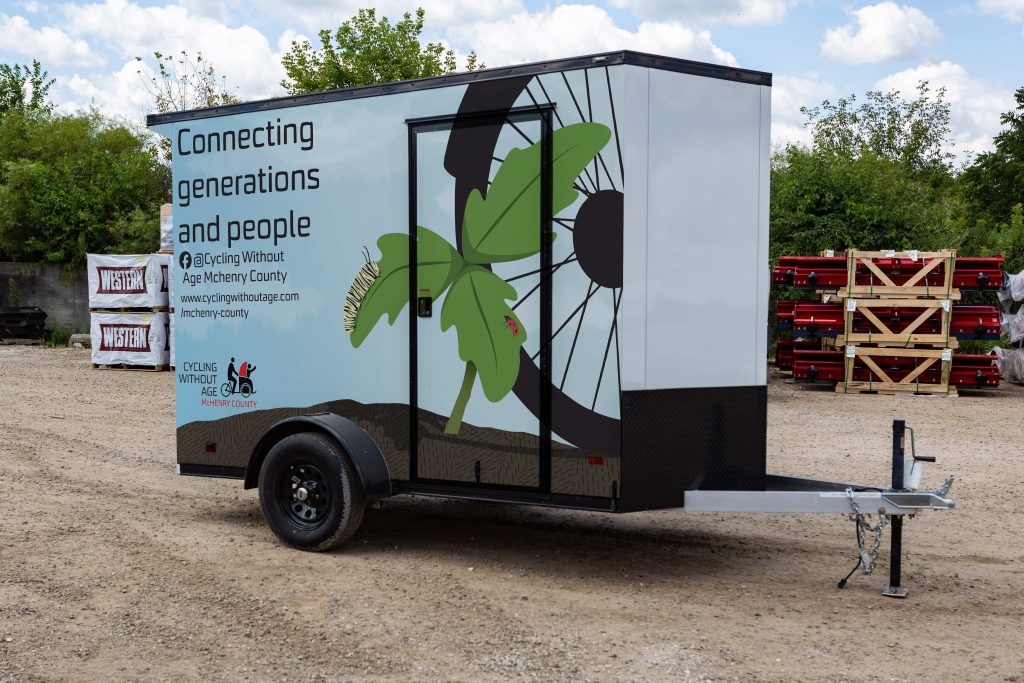 photo of a trailer with a wrap illustration depicting a sapling with a caterpillar on it and a bike tire behind it advertising cycling without age that reads "connecting generations and people"