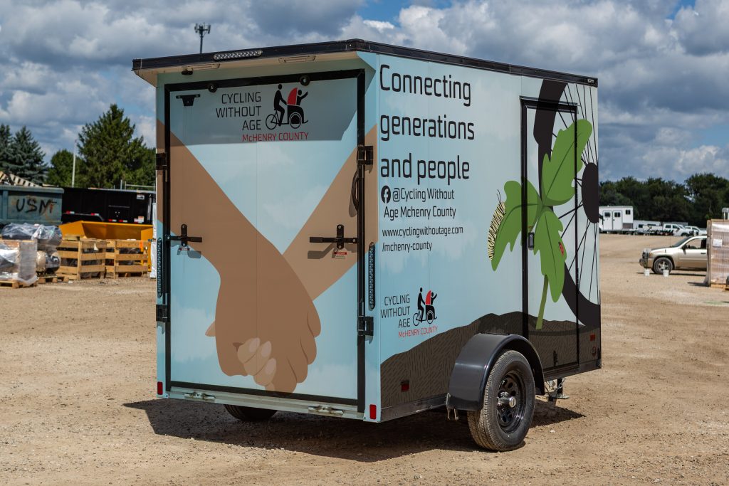 photo of a trailer with a wrap illustration depicting 2 hands holding on the back and a sapling and bike tire advertising cycling without age