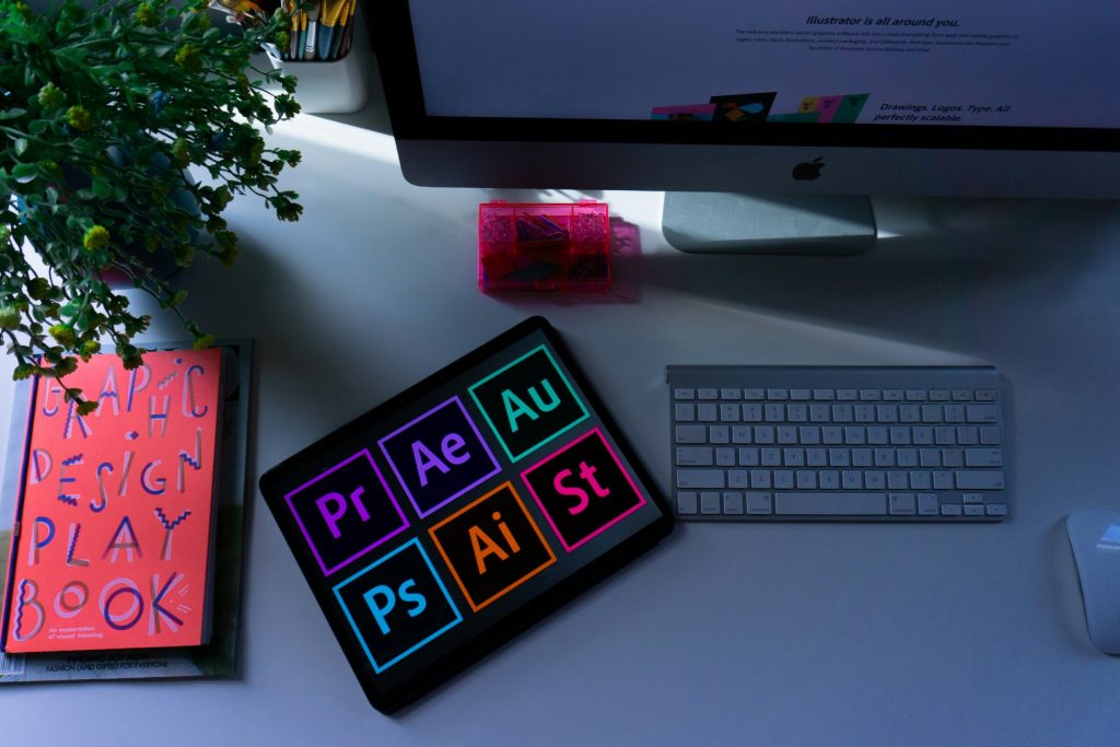 an ipad on a table displaying the logos for design programs. on either side is a keyboard and a book that reads "Graphic Design Play Book"